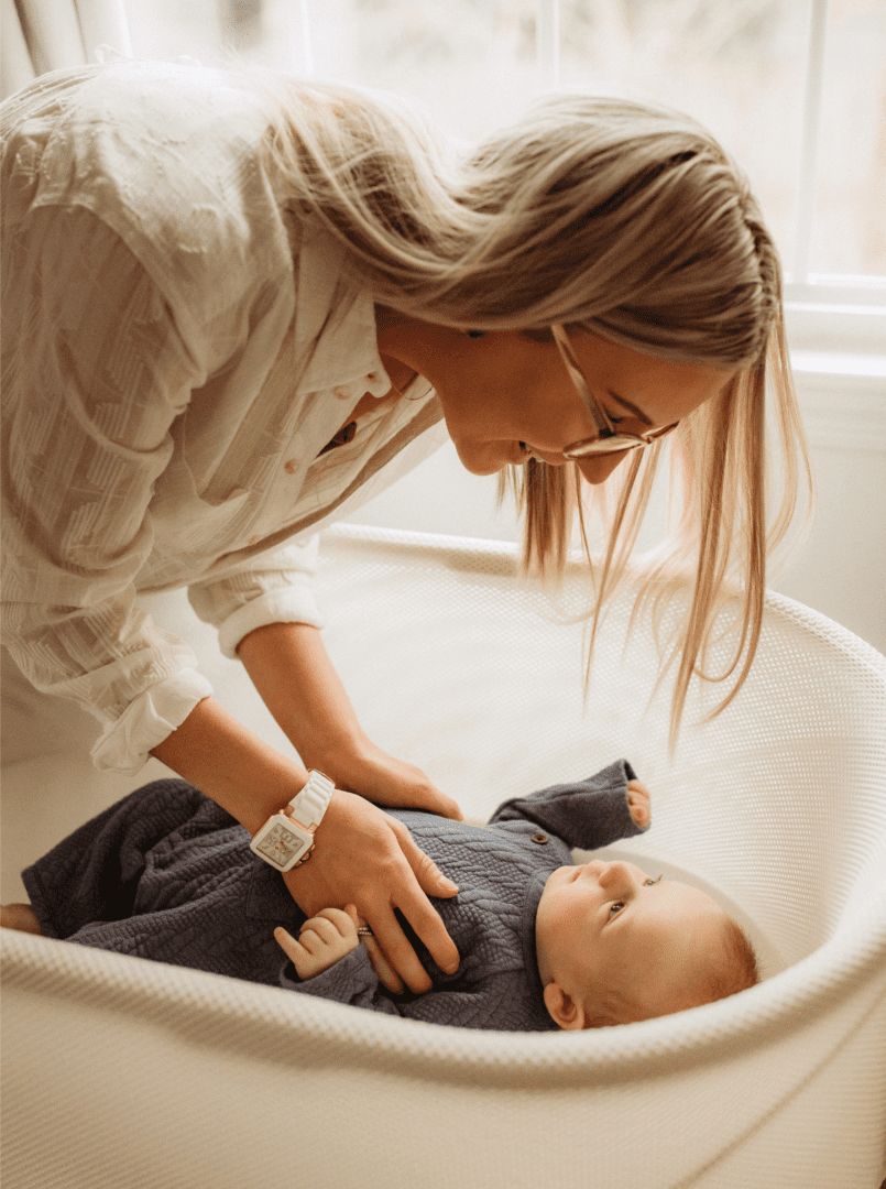 Person bending over at waist interacting with a baby laying on their back in a blue jumper in a bassinet